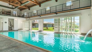 Une grande piscine intérieure donnant sur un jardin et un bâtiment en pierre en arrière-plan.
