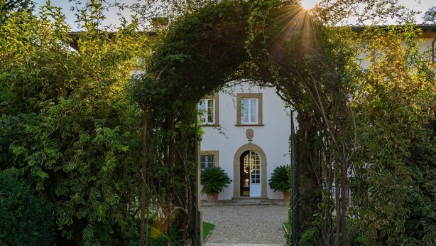 A building with a central archway surrounded by lush greenery. - Tenuta Di Pratello 24 (Peccioli)