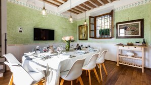 A dining area with a long table set for six, white chairs, a vase of flowers, and a television mounted on the wall. - Torre Angelina 10 2 (Montecatini Terme)