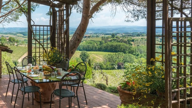 Une table dressée à l’extérieur, entourée de chaises, d’une pergola et de plantes en pot, avec vue sur un vignoble et des collines lointaines.