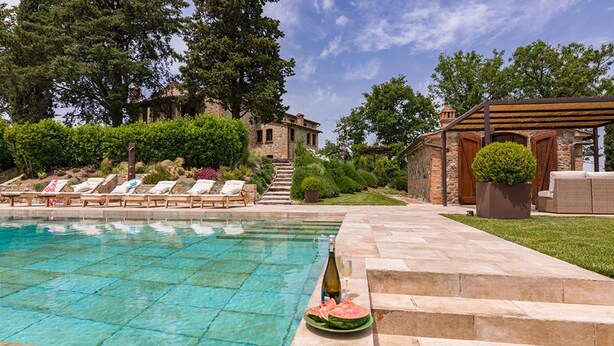 A swimming pool with lounge chairs, a stone building, and a pergola with seating.