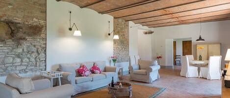 Amplia sala de estar con pared de piedra, techo de madera y zona de comedor con mobiliario blanco.