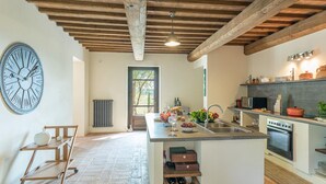 Cozinha com ilha central, vigas de madeira, um relógio grande e vista pela janela.