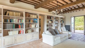 Uma sala de estar ampla com um sofá branco grande, várias estantes cheias de livros, um candelabro e vista para o verde através de uma porta aberta.