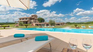 A pool area with lounge chairs and a table, a large building in the background, and a clear sky. - Podere Torricella 16 4 (Narni)