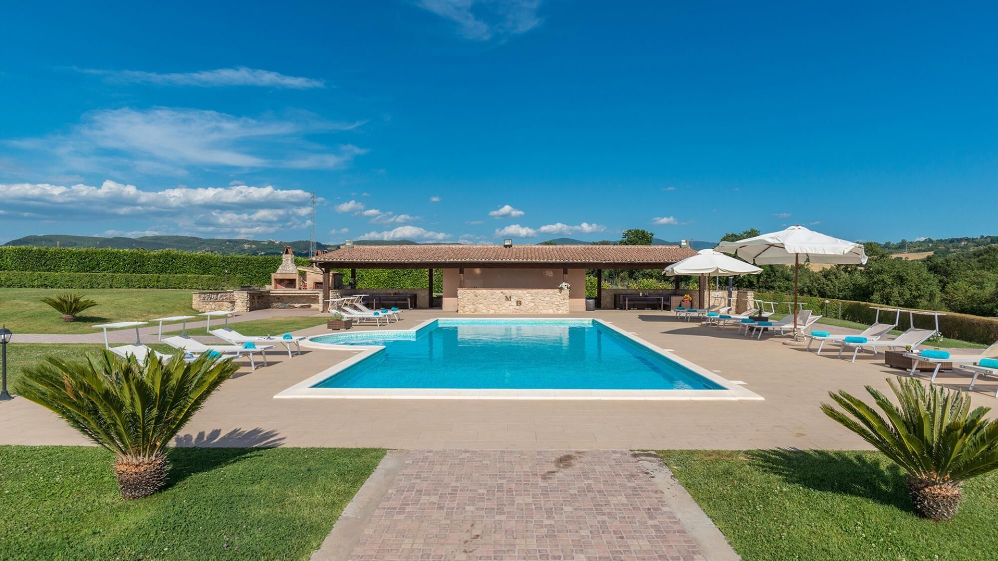 Une piscine rectangulaire entourée de chaises longues et de parasols, bordée d’un chemin en briques et de verdure, avec un bâtiment visible au loin.