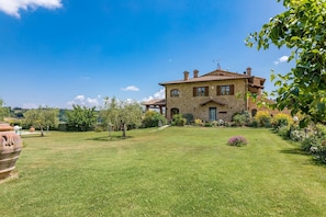 Uma casa de pedra com um gramado e jardim bem cuidados, rodeada por árvores e colinas.
