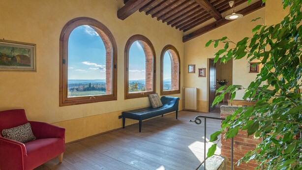 A spacious room with a wooden floor, a red sofa, a blue bench, and three large arched windows offering a view of the countryside. - Villa Siena 10 in Castelnuovo Berardenga (Castelnuovo Berardenga)