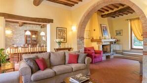 A cozy living room with a stone fireplace, a dining area with a stone wall, and a red armchair. - Villa Santa Maria 14 (Castelnuovo Berardenga)