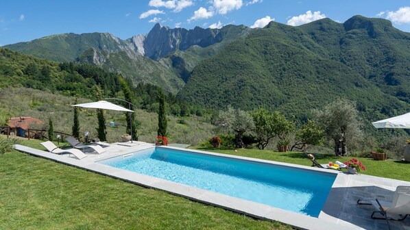 A rectangular swimming pool with lounge chairs and umbrellas, surrounded by lush greenery and mountains in the background.