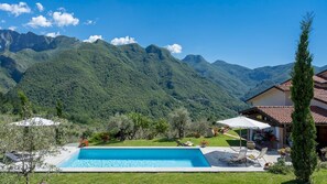 Una piscina con montañas al fondo, un césped con sillas de descanso y un edificio con tejado de baldosas.