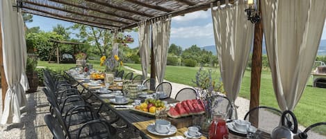 An outdoor dining area with a long table set for a meal, surrounded by chairs, with a view of a garden and mountains in the background.