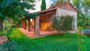 A cozy outdoor patio with a brick fireplace, a wooden table, and a comfortable seating area surrounded by lush greenery and a stone wall. - Podere Bellaria 18 2 (Asciano)