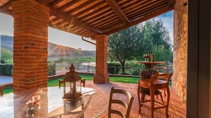 A covered outdoor patio with brick pillars, a round table, chairs, and a wine bottle and glass set. - Podere Bellaria 18 2 (Asciano)