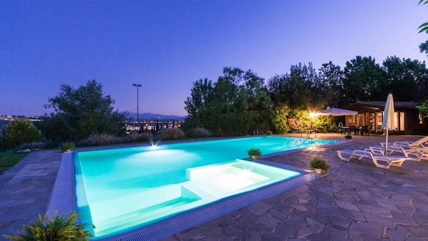 Una piscina con vistas a un paisaje urbano al atardecer, rodeada de árboles y mobiliario de exterior.