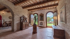 Amplia habitación con vigas de madera a la vista, pared de ladrillo y vistas a un jardín a través de ventanas de arco.