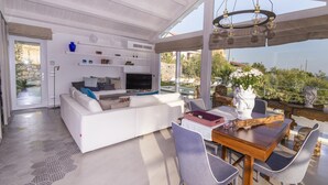 A modern living room with a white sofa, a dining table with chairs, and a TV. - Villa Aura 6 in Giardini Naxos (Giardini Naxos)