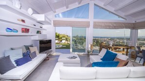 A modern living room with a large TV, white sofas, and a glass door leading to a balcony with a city view. - Villa Aura 6 in Giardini Naxos (Giardini Naxos)