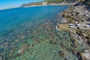 A coastal area with clear blue water, rocky shore, and a concrete structure with lounge chairs.