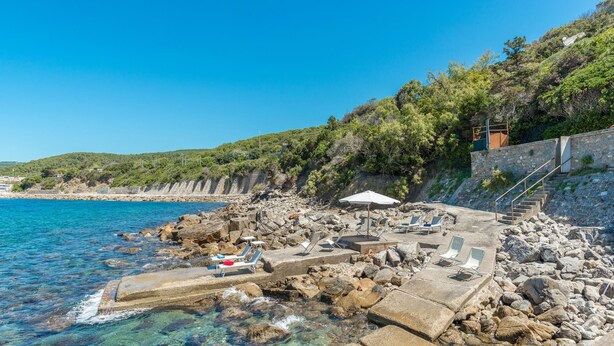 A rocky coastal area with clear blue water, a stone staircase, and lounge chairs with umbrellas. - Villa Dell Angelo 10 (Rosignano Marittimo)