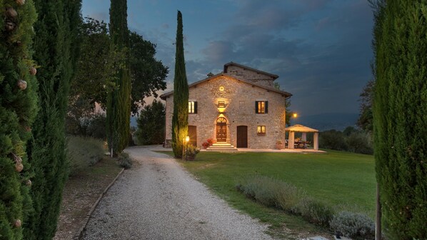 A stone house with a gravel driveway, surrounded by tall trees and a well-maintained lawn.