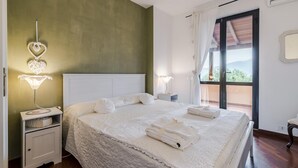 Una cama bien hecha con sábanas y toallas blancas, una lámpara de noche y vistas al exterior a través de un ventanal grande.