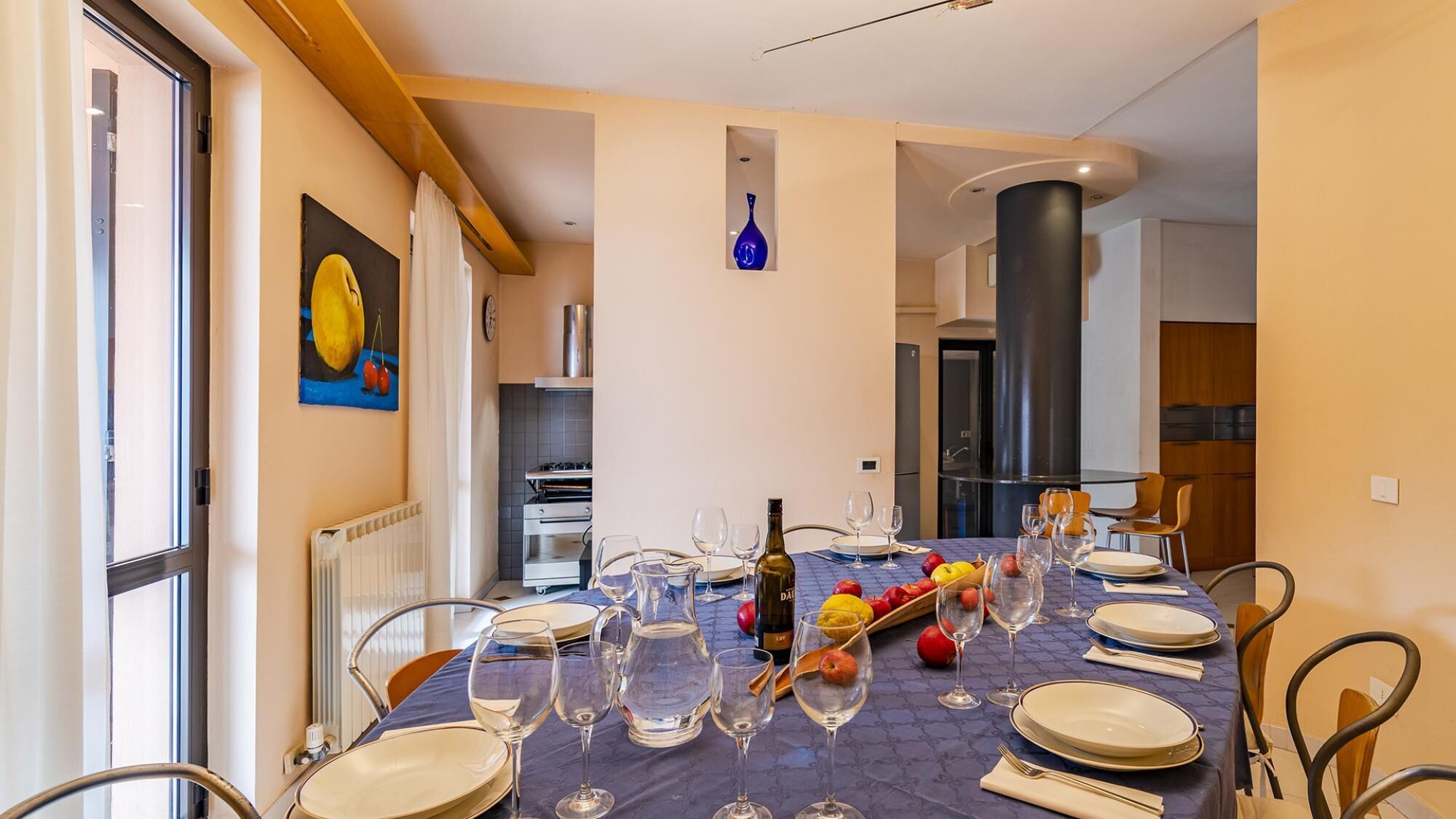 A dining area with a table set for six, including wine glasses, plates, and a bottle. There is a painting on the wall and a kitchen in the background.