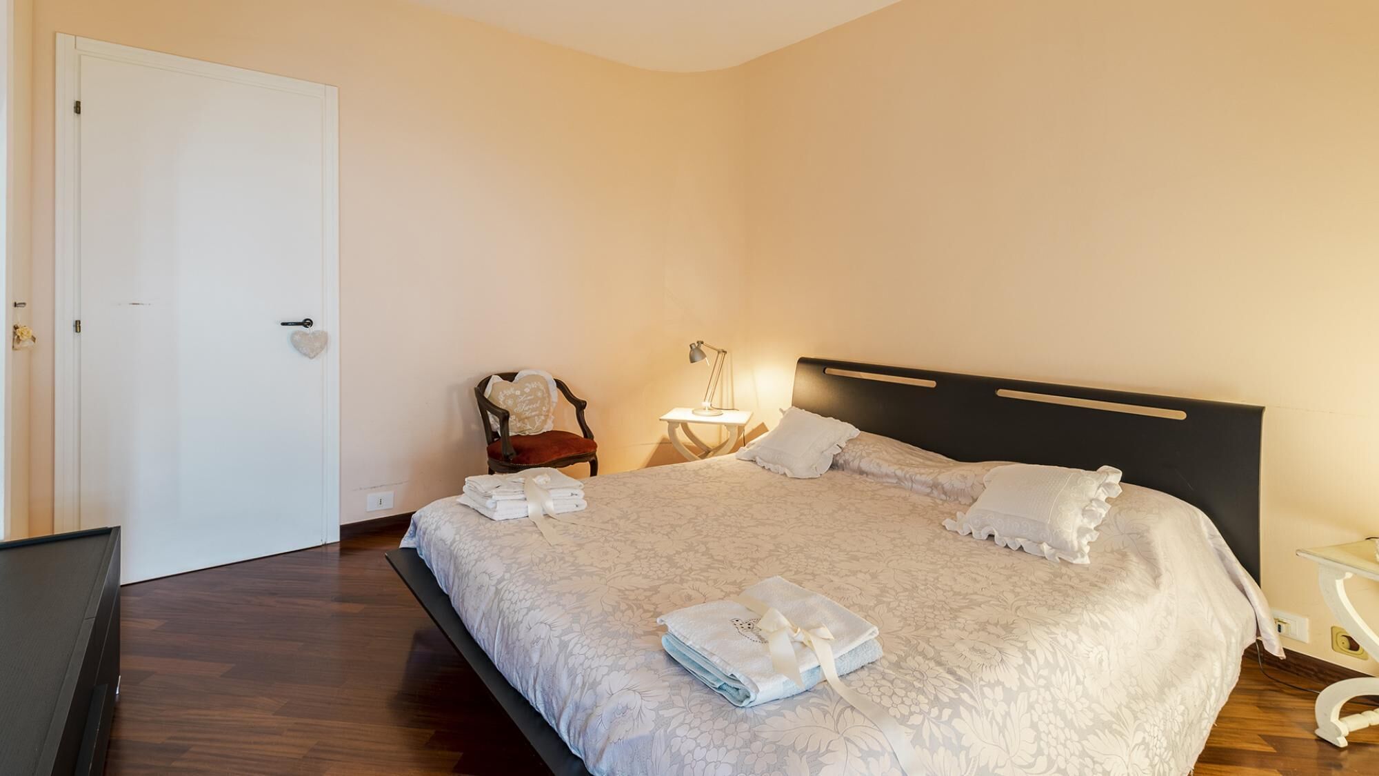 Un lit bien fait, avec des oreillers et une serviette pliée, une lampe de chevet et une chaise avec un coussin.