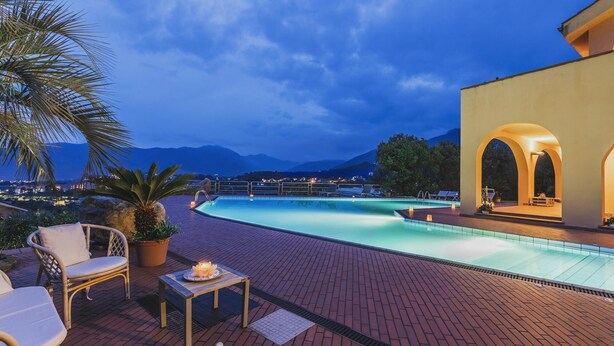 Zona de piscina con sillones reclinables y una mesa, rodeada por un patio de ladrillo y vistas a montañas.