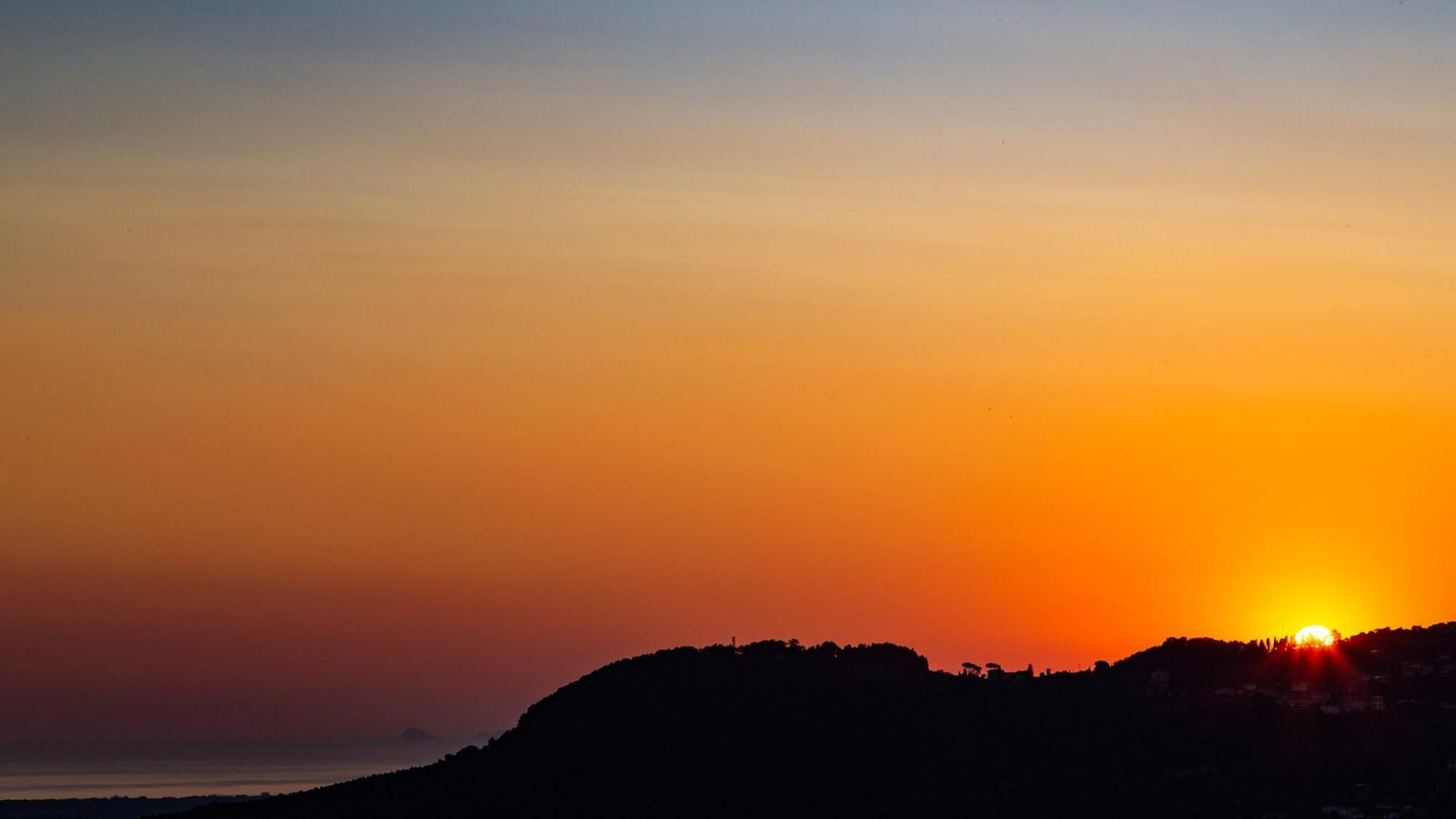 A sunset over a coastal landscape with a silhouette of a hill and a body of water in the foreground.