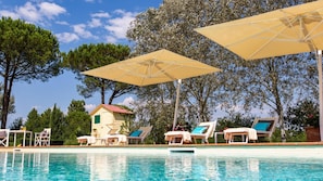 A poolside area with lounge chairs and umbrellas, a small building, and trees in the background.