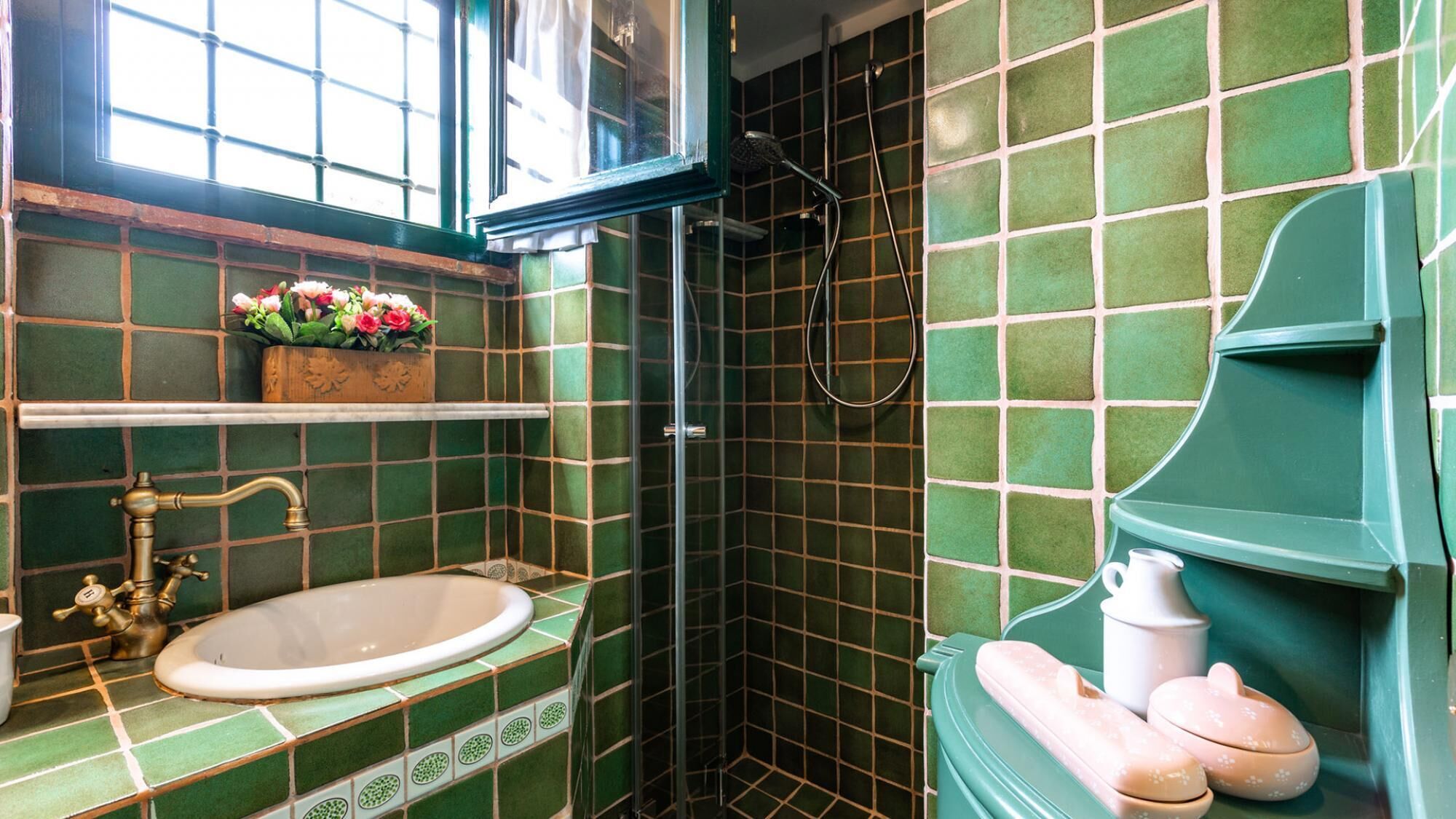 A bathroom with a green tiled wall, a white sink, a shower, and a toilet with pink slippers beside it.