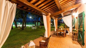 Un espace extérieur aménagé en patio, avec une pergola en bois, une table à manger entourée de chaises, et une vue sur un court de tennis en arrière-plan.