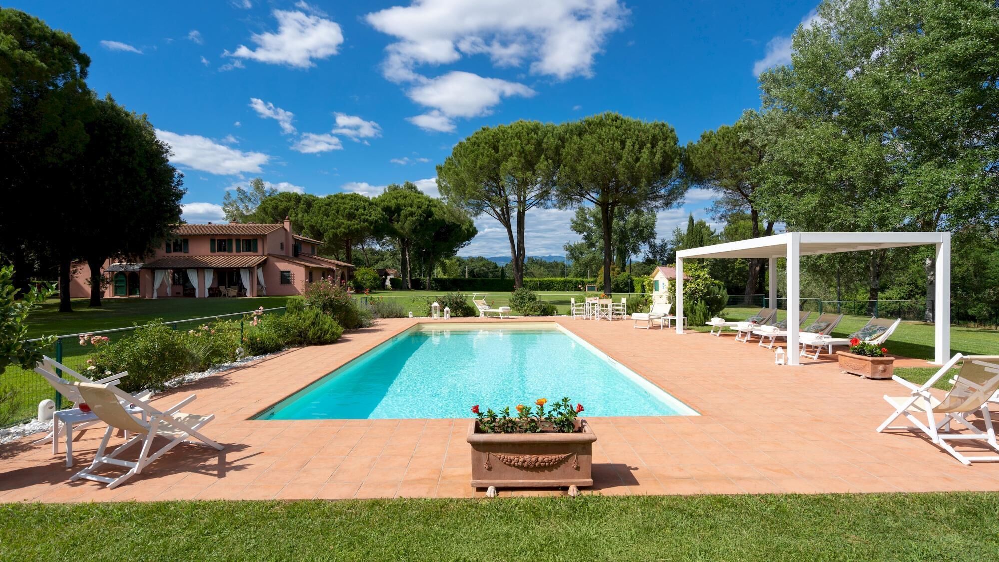 A rectangular swimming pool with a tiled deck, surrounded by white lounge chairs and a pergola.