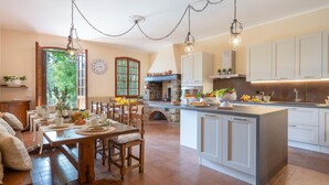 Een ruime keuken met een centraal eiland, een houten eettafel en stoelen. Er is een groot raam met uitzicht op groen.