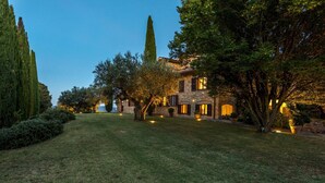 Uma villa de pedra com um gramado bem cuidado, ciprestes altos e iluminação acolhedora.