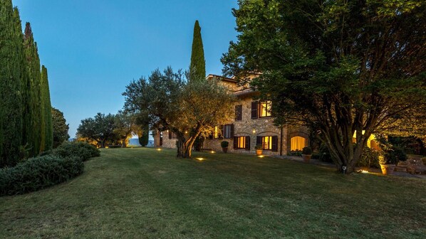 A stone villa with a well-maintained lawn, tall cypress trees, and warm lighting.