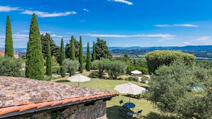 Uma villa com telhado de pedra, um jardim com ciprestes altos e vista para o campo.