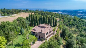 Une villa en pierre, dotée d’un toit en tuiles, entourée d’une végétation luxuriante et de grands arbres, située dans un paysage vallonné.
