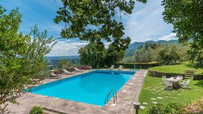 Ein rechteckiger Swimmingpool mit Steinrand, umgeben von Liegestühlen und einem Tisch mit Stühlen, vor einer Kulisse aus Bäumen und einer entfernten Bergkette.