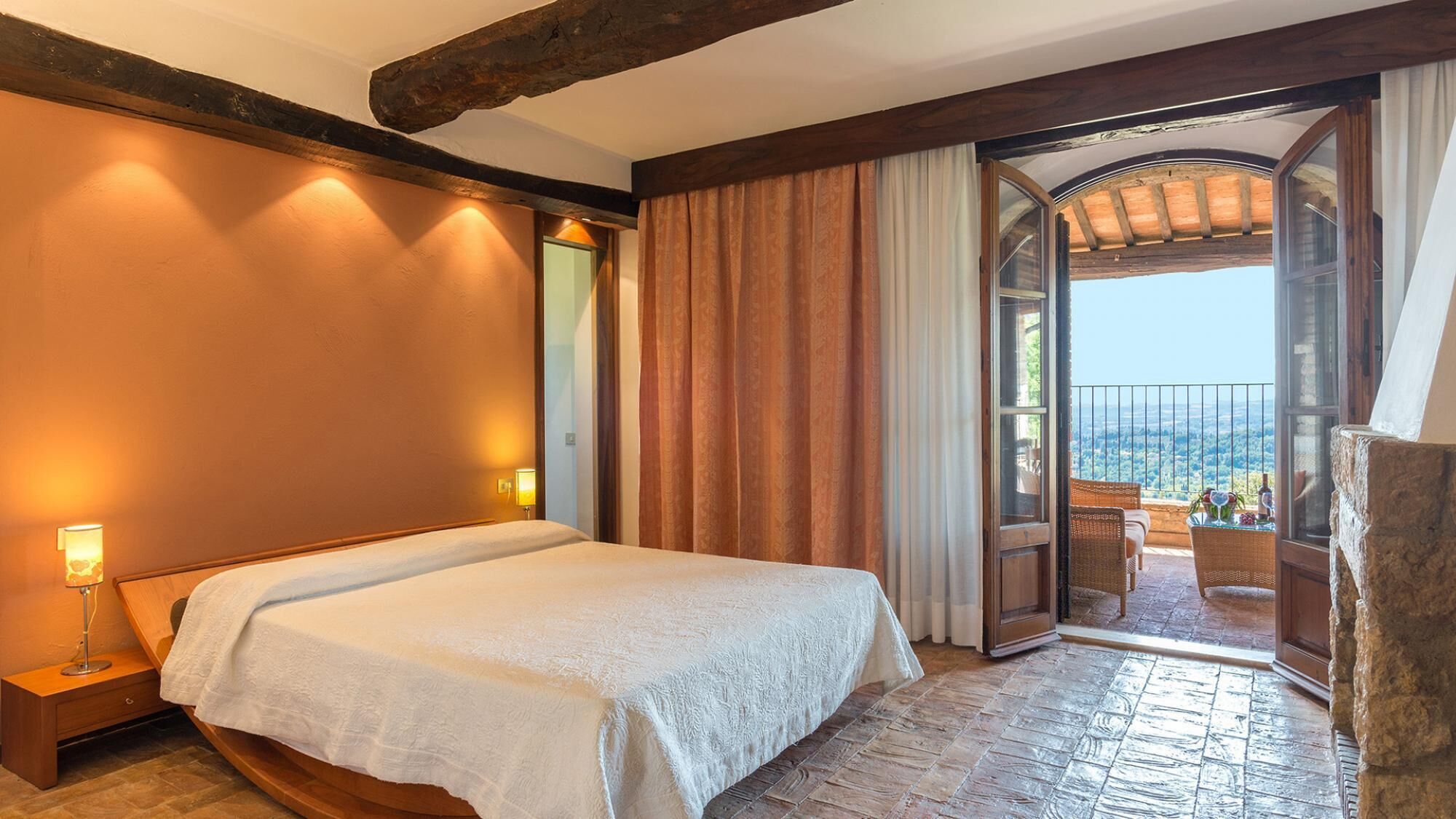 A bedroom with a large bed, a nightstand, a window with curtains, and a balcony with a view.
