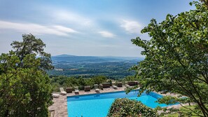 Ein rechteckiger Pool mit Steinfliesen, umgeben von Liegestühlen und einer Steinmauer, vor einer Berglandschaft und dichtem Grün.