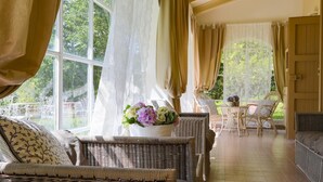 Una habitación iluminada por el sol con muebles de mimbre, una mesa con un jarrón de flores y ventanas amplias con cortinas.