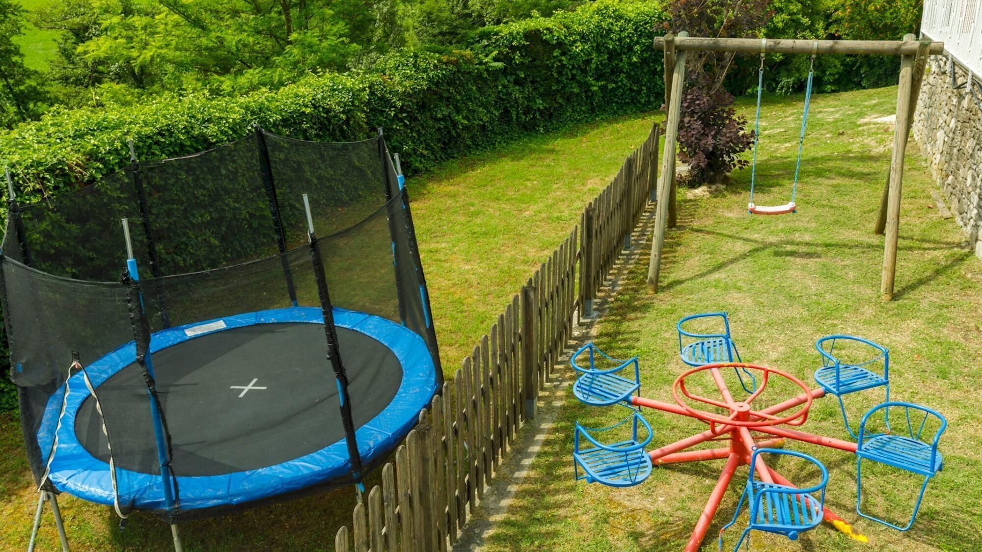 A trampoline surrounded by a wooden fence, a swing set, and a circular table with chairs in a backyard.