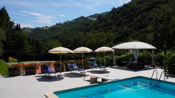 Zona de piscina con sillones de descanso y sombrillas, rodeada de un paisaje verde exuberante y colinas.
