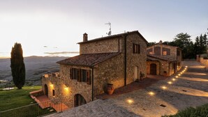 A stone building with a tiled roof, a satellite dish, and a small tower. There are potted plants and a paved pathway leading to the entrance. - La Vecchia Fonte 15 (Volterra)