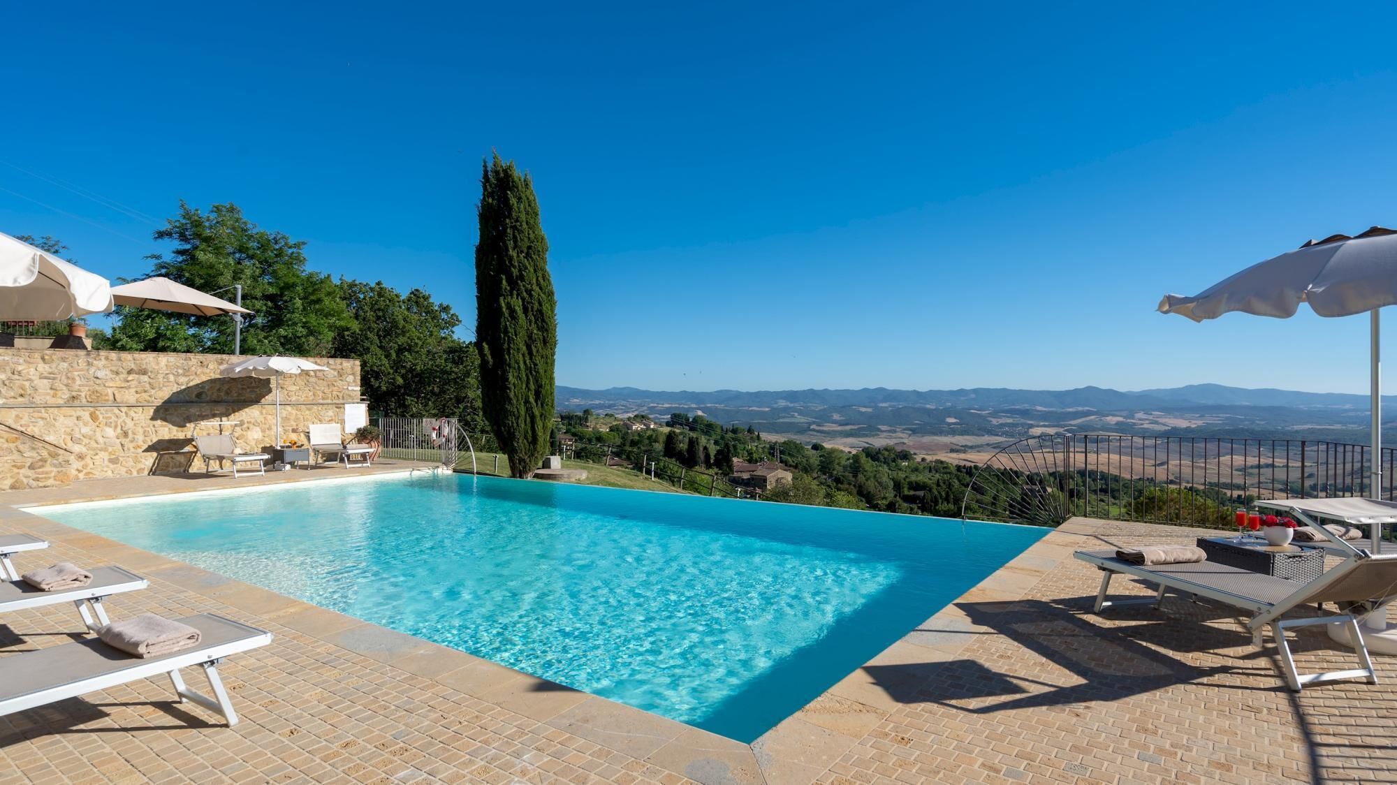 Una piscina cristallina con lettini e ombrelloni, circondata da mura in pietra e con vista su dolci colline.