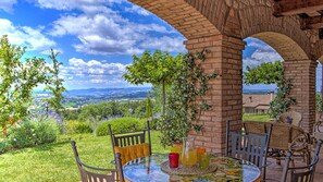 A patio with a table set for two, surrounded by wicker chairs, under a brick archway overlooking a scenic landscape.