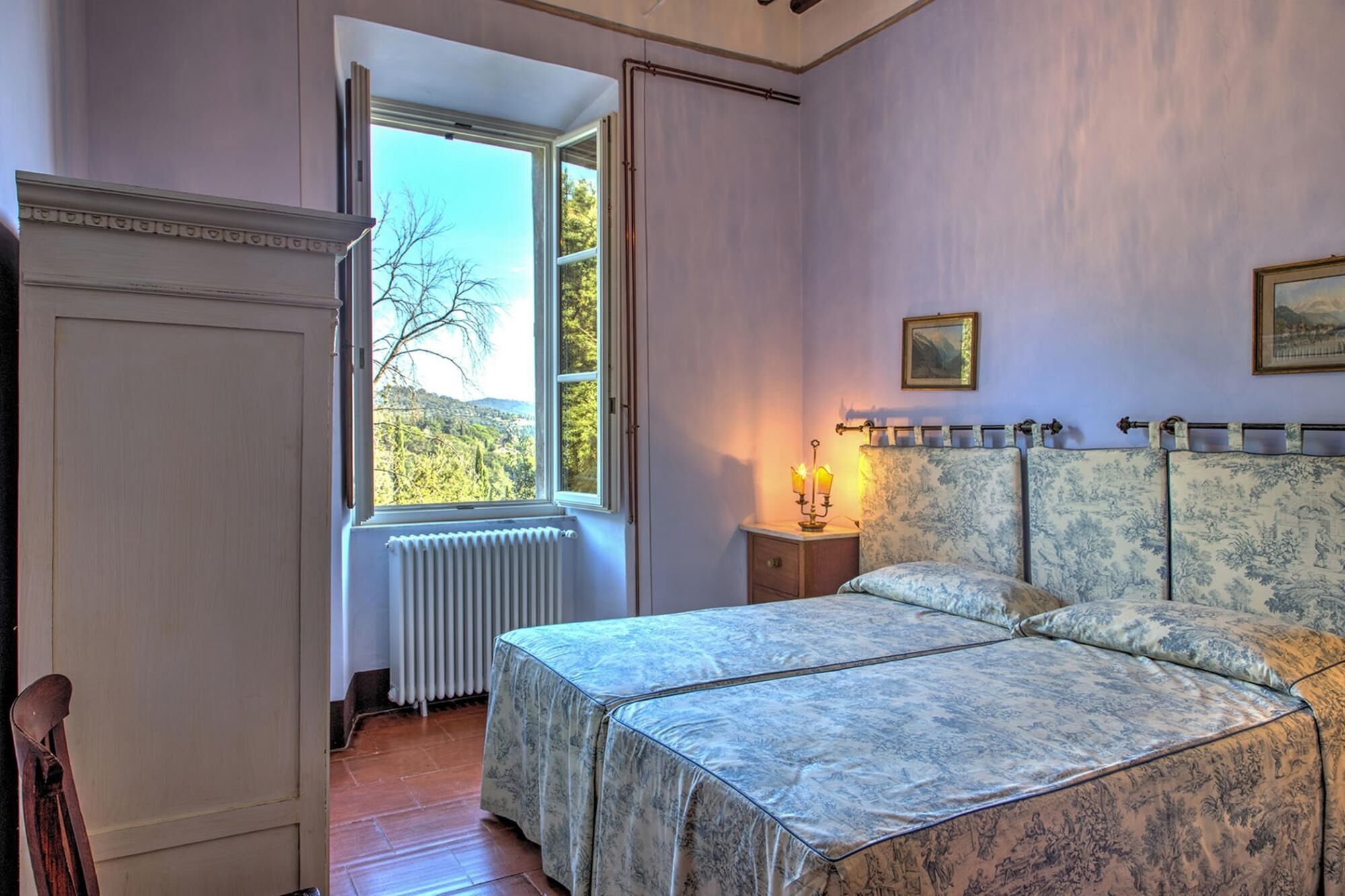 Ein Schlafzimmer mit Bett, Nachttisch, Heizkörper, Fenster mit Blick auf Bäume und einer Tür.