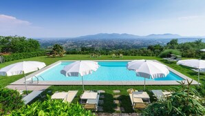 A rectangular swimming pool with white umbrellas, surrounded by lounge chairs and greenery, with a view of a city and mountains in the background. - Villa Primo Sole 22 (Lucca)
